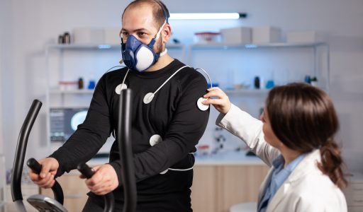 Man athlete with mask running on cross trainer after medical researcher attached electrodes to her body. Physician using computer to controls EKG data showing on laboratory monitors, VO2 max test.